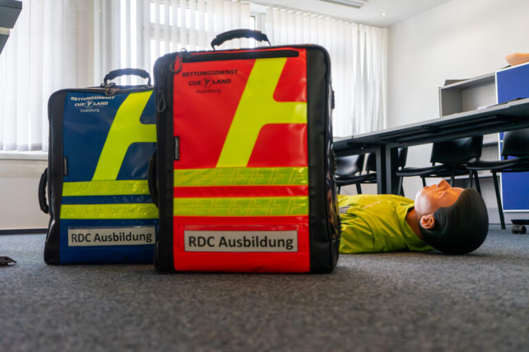 Zwei Notfallrucksäcke aus dem Bildungszentrum der RDC stehen auf dem Boden vor einer Reanimationspuppe